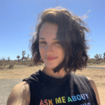 Izzy has chin-length brown hair and is smiling at the camera. She is in the desert on a sunny day wearing a black tank top with rainbow lettering.